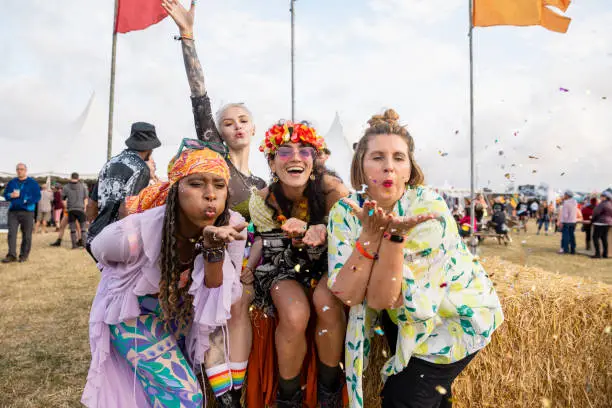 Group photo of four female friends blowing confetti into the camera lens. They are all dressed in vibrant colours and clothing at a festival in Northumberland in the north east of England.