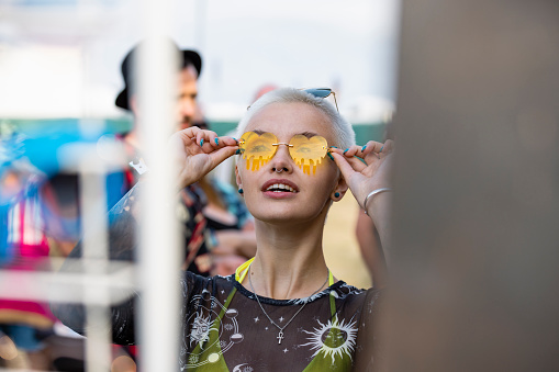 A portrait of a young woman with short hair trying on a pair of sunglasses at a summer festival. She is looking up and smiling with joy as she places the sunglasses on her face. The festival is in northumberland in the north east of England.