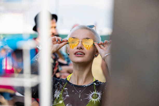 Festival Sunglasses A portrait of a young woman with short hair trying on a pair of sunglasses at a summer festival. She is looking up and smiling with joy as she places the sunglasses on her face. The festival is in northumberland in the north east of England. festival goer stock pictures, royalty-free photos & images