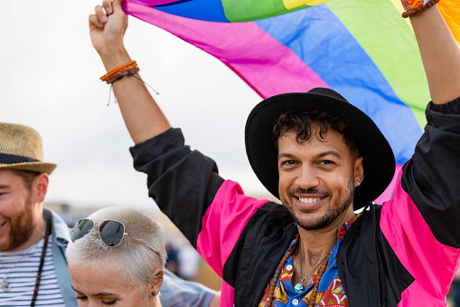 A photo of a young man at a festival waving a stripy multicoloured flag above his head. He is wearing a black fedora hat and he is smiling and having a fun.