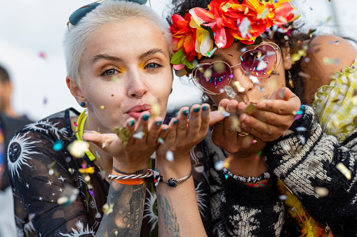 A photo of two young woman at a festival blowing confetti  into the camera lens. One of the women is wearing sunglasses and a floral crown on her head.