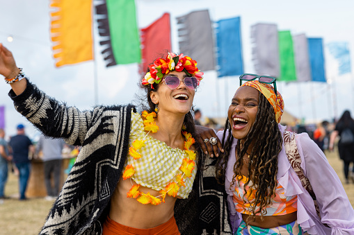 Two female friends laughing and embracing each other at a festival. They are both laughing and smiling hysterically as they walk. They are both dressed colourfully.