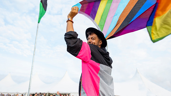 A shot of a young man smiling and waving a flag above his head at a festival. The festival is in northumberland in the north east of England. He is wearing a vibrant jacket and a black fedora hat on his head.