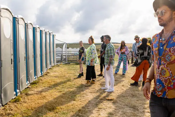 A group of people standing outside on a sunny day and queuing in line for the portable toilet at a music festival event in the north East of England.