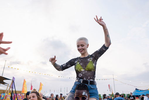 A young blonde woman with short hair sits on her friends shoulders at festival as she embraces the atmosphere. She has her hands in the air as she enjoys the moment.