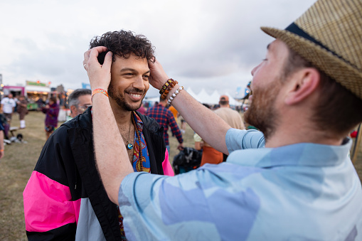A shot of a gay male couple interacting with each other at a festival. One of them is styling the others hair. He is wearing a fedora hat and a blue shirt.