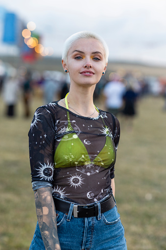 A waist up portrait shot of a young woman at a festival. She is looking into the lens and smiling pleasantly. She has short blonde hair and is wearing a black shirt and shorts.