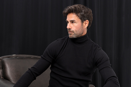 Middle aged handsome Hispanic man wearing black turtleneck sweater sitting on leather sofa in front of black curtain and looking away.