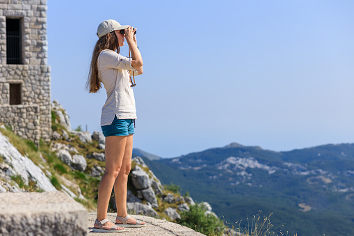 Young woman watching mountain landscape through binoculars at the view spot on tourist trail.