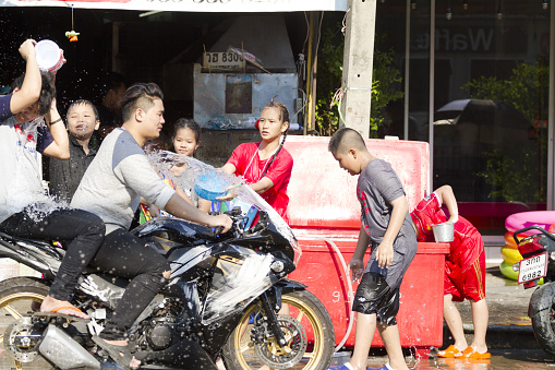 Two thai men on motorcycle are getting water over body while driving slowly along street Chokchai 4 in Bangkok Ladprao at buddhist new year and water Songkran festival. In background are teenagers