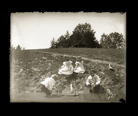 Beautiful Black and White portrait of four Victorian-era children playing with a toy boat in a pond wearing period clothing. The image was digitally restored from a glass plate taken circa 1890.