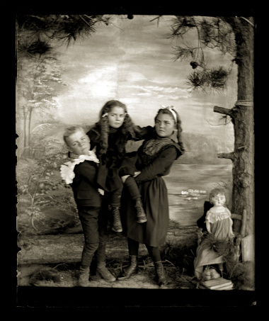 Beautiful Black and White portrait of three Victorian-era children playing wearing period clothing posed in front of a painted backdrop. The backdrop was hand painted and placed on the side of a house to create the illusion of a studio space. The image was digitally restored from a glass plate taken circa 1890