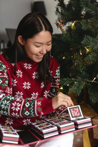 jeune femme asiatique ouvrant le calendrier de l’avent assise près du sapin de noël dans le salon - advent calendar advent christmas tree christmas photos et images de collection