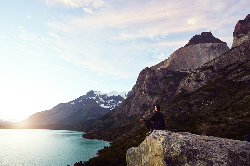 silhouette of a man sitting meditating on a rock over the lake with mountains and trees in the background in Paine Grande of Torres del Paine National Park