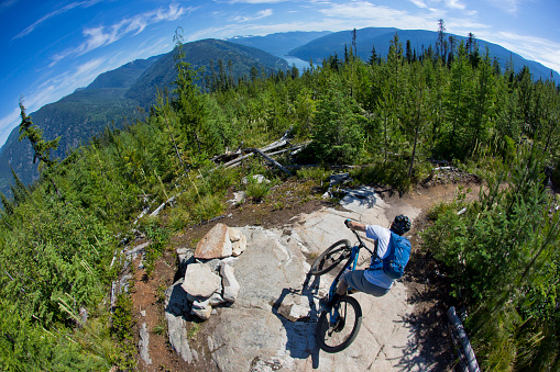 A man goes for a mountain bike ride near Nelson, British Columbia, Canada. He is riding a cross-country style mountain bike and wears a backpack and cycling helmet. Kootenay Lake is visible in the background.