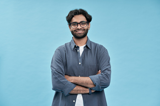 Smiling confident arab young man, male student, professional employee or programmer standing isolated on blue background. Happy handsome ethnic guy wearing shirt and glasses portrait.