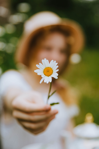 a girl in a hat on a flowery field covers her face with a daisy