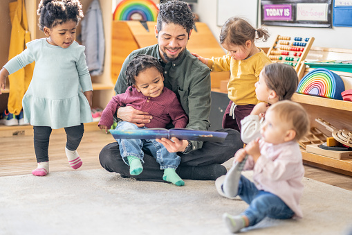 A male daycare educator of Hispanic decent, sits on the floor with a child on his lap and other toddlers scattered around him, as he reads them a story.  They are each dressed casually and are focused on the book.