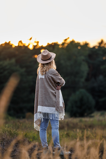 Woman wearing poncho and cowboy hat and enjoying sunset in nature. Female person wearing boho fashion style