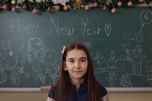 Portrait of a little cute girl standing in the classroom in front of a chalkboard and photographing for a New Year school event.