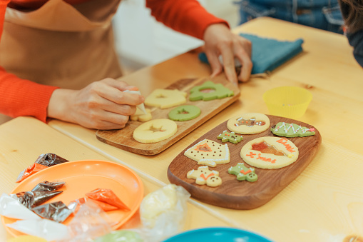 Close up of Asian woman female hands placing baked and decorated Christmas cookies on  wooden board ready to serve, with other cooking utensils in kitchen.

Moments of young Asian family members, parents and children spending time and having fun together during Christmas winter holidays.