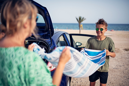 Mother and teenage kids returning to car from the beach. Family is packing the car. Teenage boy and his mother are shaking out a towel from the sand.\nSunny summer day in Spain.\nNikon D850