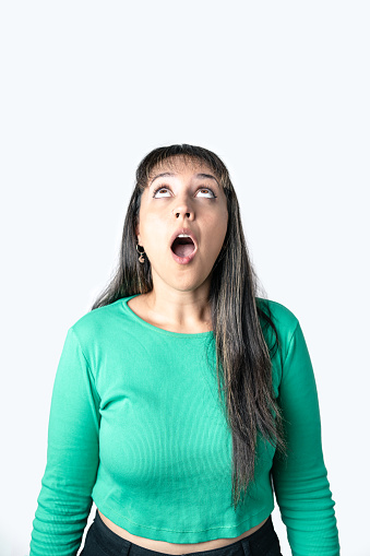 Portrait of surprised Latin woman on green blouse opening her mouth and eyes and looking up at something. White isolated background for copy space