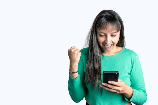 Happy Hispanic woman dressed in casual clothes looking at her smart phone screen and celebrating on white isolated background stock photo