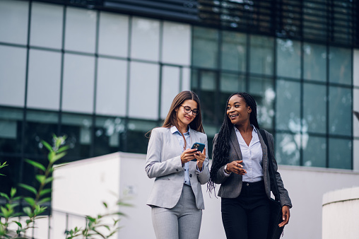 Team of multiracial successful business women on their way to the office talking about strategy and using a smartphone. Diverse women walking outside with buildings in the background. Teamwork.