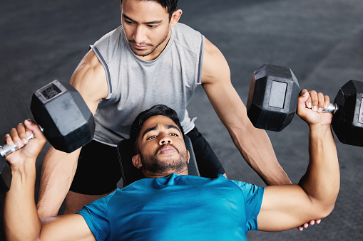Fitness, dumbbell training and personal trainer men with support, trust and muscle help in gym together. Helping, coaching and motivation of coach for man bodybuilder challenge, exercise or workout