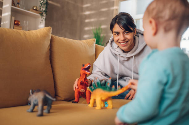 Mother and toddler son playing with dinosaurs toys at home on the couch stock photo