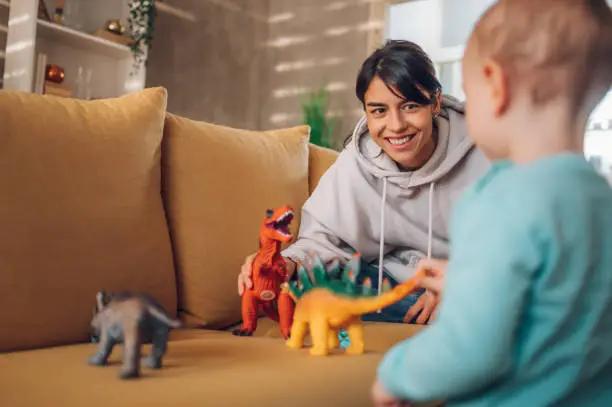 Young mother and toddler child enjoying developing activities at home while playing with dinosaurs toys on a couch. Focus on a mother. Educational games, having fun with kid at home. Copy space.