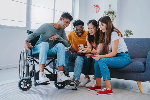Man on wheelchair with friends having fun watching smart mobile phone device - Happy teenagers posting picture on social media - Youth lifestyle, healthcare and  institutions for the disabled concept
