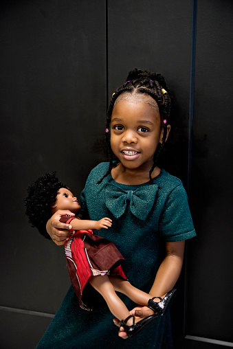 Portrait of black little girl with same skin colour doll. She is six year’s old and is looking at the camera. Vertical waist up indoors shot with copy space and black background. This was taken in Montreal, Quebec, Canada.