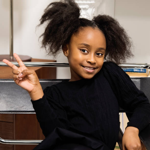 Portrait of black little girl making a peace hand sign. Portrait of black little girl making a peace hand sign. She is eight year’s old and is looking at the camera. Square waist up indoors shot with copy space and living room background. This was taken in Montreal, Quebec, Canada. little black girl hairstyle stock pictures, royalty-free photos & images