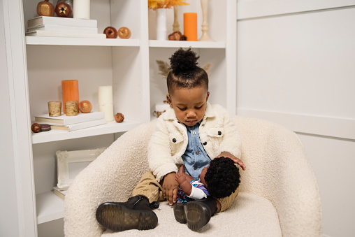 Portrait of black little boy with same skin colour doll. He is 18 months and is looking at the doll. Horizontal full length indoors shot with copy space and living room background. This was taken in Montreal, Quebec, Canada.