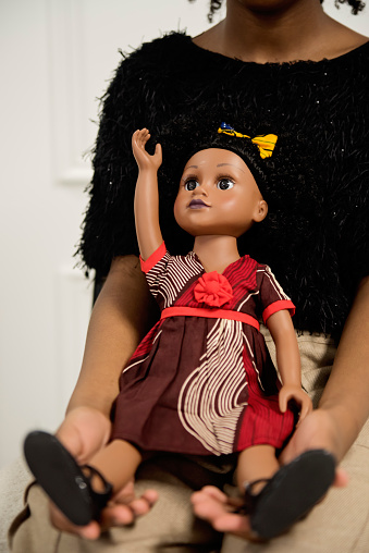 Close-up of black little girl with same skin colour doll. She is ten year’s old and is holding her doll. Vertical waist up indoors shot with copy space and white background. No face. This was taken in Montreal, Quebec, Canada.