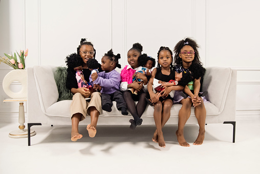 Black little girls sitting on a couch with same skin colour dolls. They are from three to nine year’s old and are looking at the camera. Horizontal full length indoors shot with copy space and white background. This was taken in Montreal, Quebec, Canada.