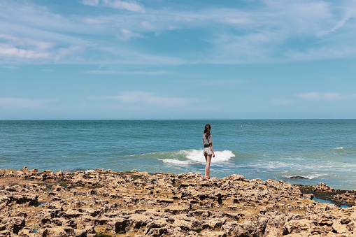 Caucasian teenager brunette girl in shorts and a blouse from the back stands on a rocky shore looking at the north sea in Etretat in France, close-up side view. People and nature concept.