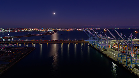 Drone shot of the harbor in the Port of Los Angeles, looking past rows of container cranes towards Navy Way. A bright full moon is shining down, reflecting in the water. \n\nAuthorization was obtained from the Los Angeles Harbor Department and Los Angeles Port Police for this operation in restricted airspace.