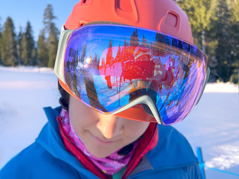 Eurasian young adult daughter snowboarder standing in short ski lift lineup.  POV - reflection in goggles shows mature Asian mother taking smartphone photo of daughter.  North Vancouver, British Columbia, Canada.