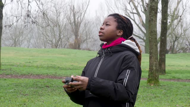 Young african girl standing checking flying 4k drone outdoors in a park in cold weather.