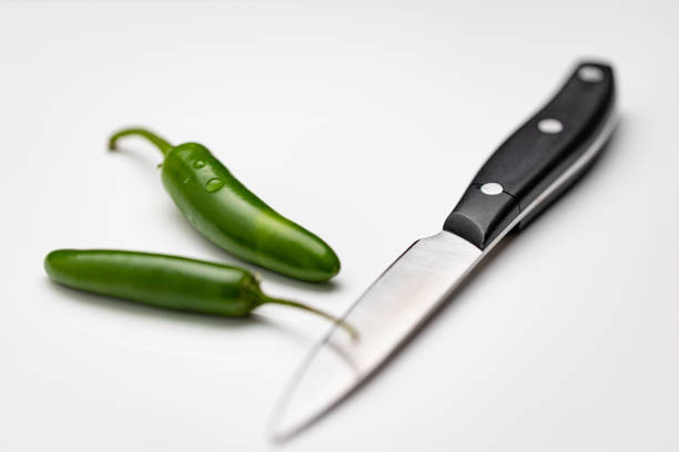 Two Jalapeños green chili peppers on white background next to kitchen knife ready for slicing