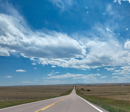 This long, straight country road goes through miles of ranchland in Colorado.  Highway photo taken on July day with white, wispy clouds. Prairie grasses, cattle and bison are seen along the road to the mountains in the distance.