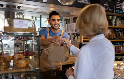 Happy Latin American waiter giving a paper bag to a client buying pastries at a cafe