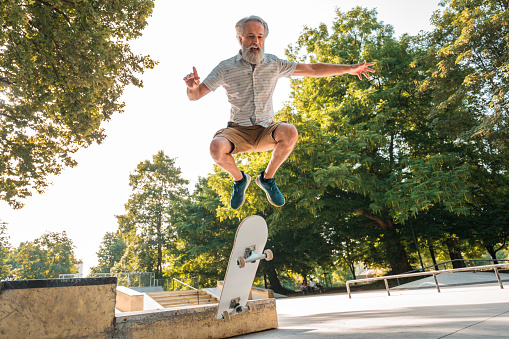 Full length low angle view of a mature Caucasian man with gray beard flying in the air with a skateboard. He is happy and enjoying a fun day outdoors.