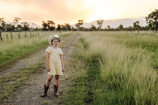 Young cowgirl standing on a on a dirt road at sunset wearing a dress, boots and a cowboy hat.