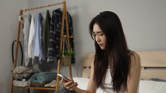 upset chinese female combing hair with hand and propping face while checking emails for her stressful work on the phone after getting up in the morning at home