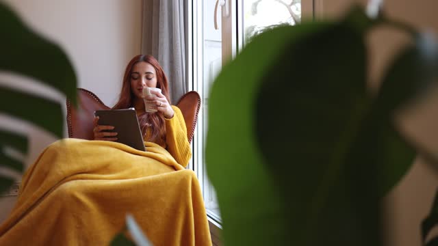Young attractive redhead woman holding a tablet at home, with a blanket in the armchair by the window.