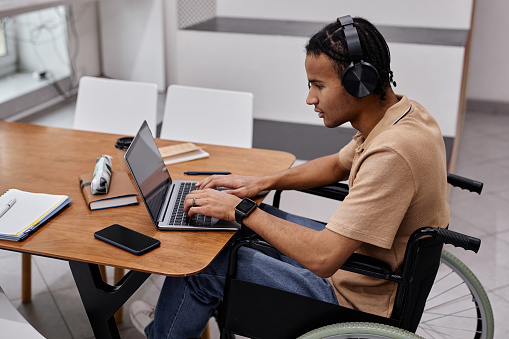 Portrait of black young man with disability using laptop in college library and wearing headphones